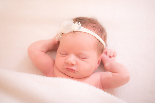 A sleeping two week old newborn baby girl wearing a vintage lace headband with flower. She is wrapped in gauzy cream colored fabric and sleeping on a white billowy blanket.