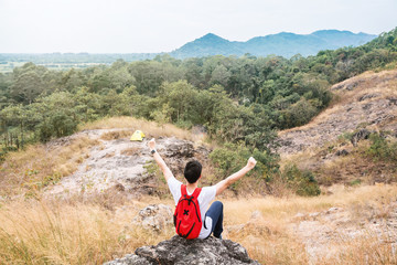 Fototapeta premium Rear view of young man backpack traveler sit on stone at peak of mountain and had rise arms up to sky. Victory and success concept.