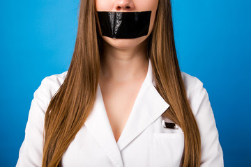 Girl doctor standing in a white coat with black tape over her mouth on a blue background close-up,...