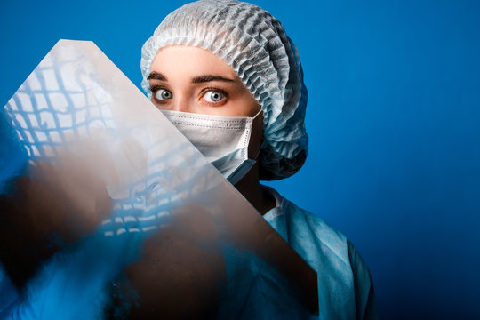 Doctor Renginologist In Blue Uniform Standing On A Blue Background In A Medical Protective Mask Holding A Chest X-ray In His Hands Closeup Peeping From Behind A Snapshot