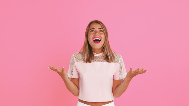 Pretty woman with shiny freckles, dressed in top. She is looking confused, gesticulating and asking why, posing against pink background. Close up