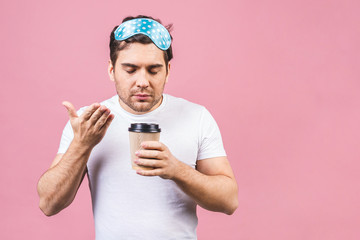 Portrait of nice calm peaceful sleepy young man with blue sleeping mask and cup of coffee or tea. Isolated over pink background.
