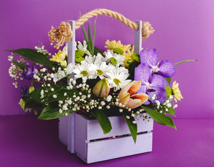 Beautiful bouquet flowers in lilac wooden basket on purple background. Orchids, chrysanthemums, lilies, chamomiles floral floristic composition. Greeting card for romantic date, valentine day, 8 March