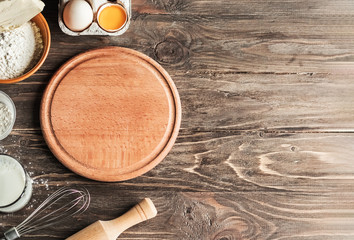 Baking cooking ingredients flour, eggs, rolling pin, butter, cottage cheese and a wooden round board on a wooden background. View from above. Copy space. Cookie pie or cake recipe layout