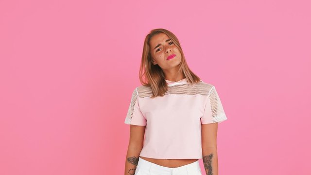 Pretty female with tattooed hands, dressed in top. She is looking disappointed, gesticulating and asking what, posing on pink background. Close up