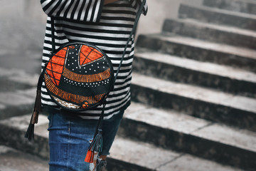 Fashionable girl holds bag dressed in a striped black white sweater on a gray background. Stylish round bag with designer pattern. Boho style. Crossbody bag. Female fashion concept. Copy space.