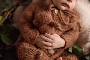 A newborn charming boy, 5 days old, sleeps in a cozy bear outfit and in a light brown outfit. He sleeps in a basket and holds a tiny teddy bear.