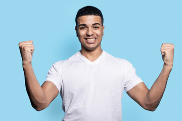 Naklejka premium Overjoyed black man raises clenched fists, feels energized while gesturing happily against blue wall
