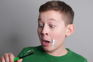teenager european boy grimaces and staring at  toothbrush and toothpaste on cheeks in green sweater on grey background