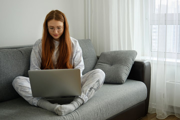 Obraz premium Red-haired girl in pajamas and glasses sits on a sofa and types on a laptop