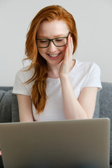 A red-haired girl sits on a sofa in a white T-shirt, smiles, adjusts her glasses and looks at the laptop.