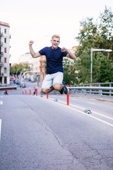 Young blonde male jumping outdoors while looking camera in the street