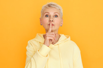 Portrait of blue eyed blonde mature woman in hoodie holding fore finger on her lips, shushing,...