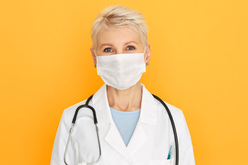 Portrait of attractive blonde senior woman doctor wearing medical coat and white surgical mask against infection, virus or bacteria during pandemic. Healthcare, prevention and protection concept
