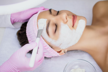 The cosmetologist applies a white clay mask to the face of a beautiful young woman with a white...