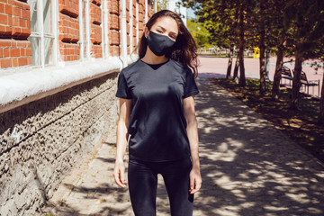 Young woman in a black t-shirt and a protective face mask stands on the street.