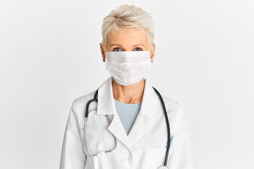 Healthcare and wellness concept. Studio image of serious short haired blonde mature woman physician posing isolated wearing uniform and white medical mask against viruses, bacteria or infection