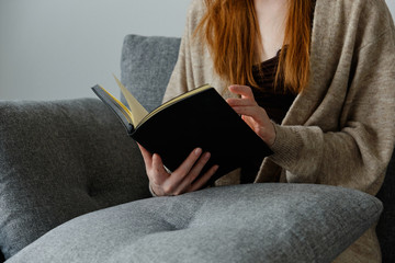 Obraz premium A black book in the hands of a red-haired girl sitting on a sofa.