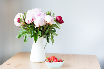 Bunch of Pink peonies in vase and strawberry on the wooden table . Flowers on a beige wooden table near the window. Home house interior. Beautiful peony flower.Summer day. Spring. Copy space
