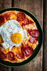 Sourdough breakfast pan pizza with pepperoni and fried egg in a green plate on wooden surface