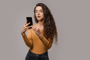 Fototapeta premium Chatting on mobile cell phone, online internet technology concept. 5g connecting and communication. Amazed emotional woman with shocked looking at phone, reaction on news over grey background