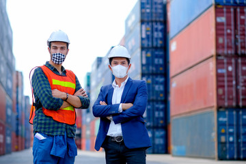 Technician or engineer workers stand in front of shipping container with one hold infrared thermometer and manager also wear face mask in the procedure to prevent virus infection in workplace area.