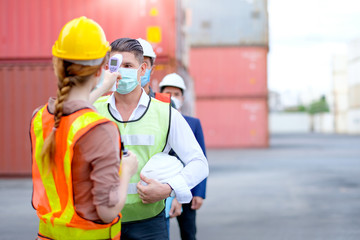 Technician or engineer worker use infrared thermometer measure temperature of other staff in shipping container workplace area and they stay in line during covid pandemic in around the world.