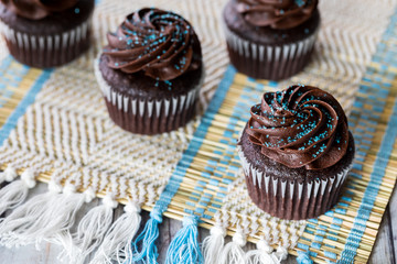 An above close up view of chocolate cupcakes with chocolate frosting and blue sprinkles on a woven matt.