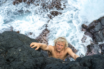 A young climber with long white hair climbs a rock against the background of the ocean