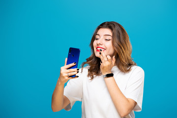Girl with a phone in her hands smiles and does makeup, isolated on a blue background.
