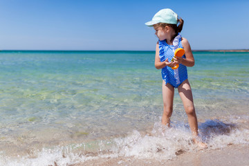 Fototapeta na wymiar Happy cute tollder girl 4 or 5 years old in a bright blue swimsuit with toys playing in the water at sea on a sunny summer day. Fun family vacation at the sea. Kids activity outdoors
