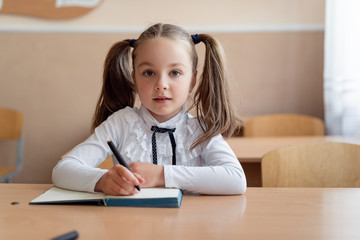 Naklejka premium little girl schoolgirl is sitting at a desk. Hairstyle two braids on the head. Hold a pen and look at the teacher. Write in a notebook.