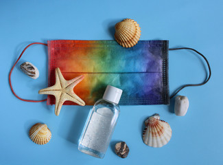 Summer vacation flat lay with sea shells, pebbles and starfish next to protective mask decorated in rainbow colors and antiseptic on blue background. Travel and coronavirus covid-19. 