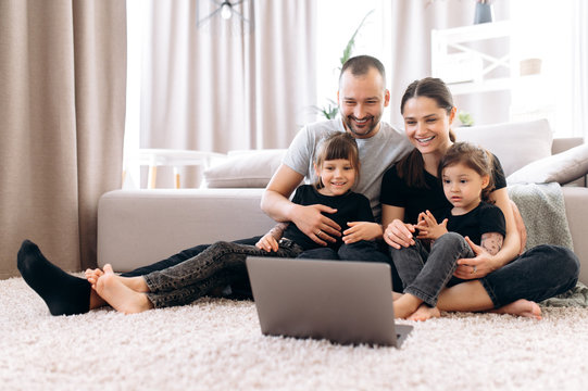 Video Conference. Big Happy Family. Dad Mom And Their Two Amazing Daughters Communicate Via Video Conference With Grandparents Sitting At Home On The Floor In A Positive Mood And In Stylish Clothes