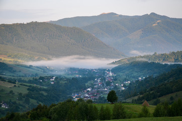 Beautiful summer landscape of the fog over small village in the valley. Golden sunrise in the morning Carpathian Mountains