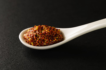 Whole grain mustard in a white chinese porcelain spoon on a black stone serving board. Sauces and seasonings for meat, fish and vegetables.
