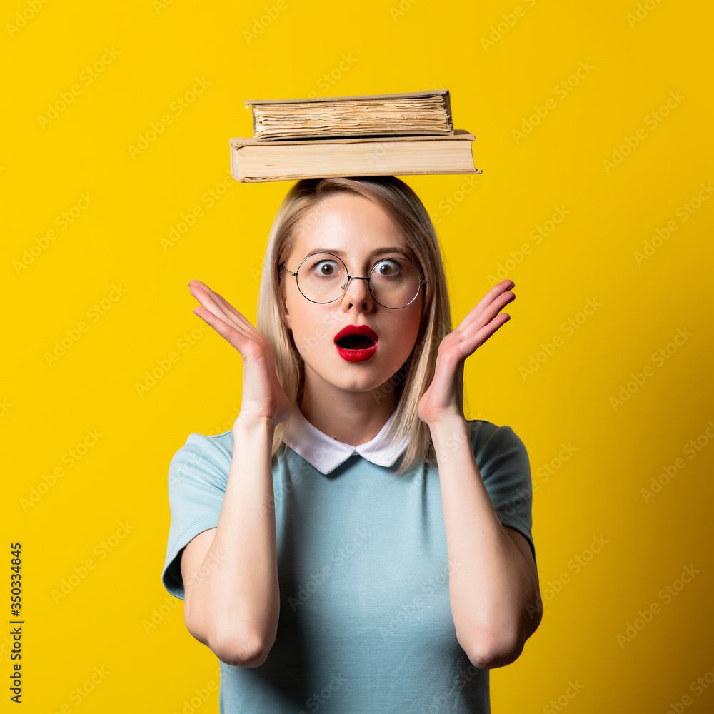 Wall mural Blonde girl in blue dress and glasses with books on yellow background