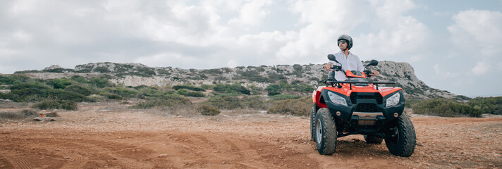 A man sits on a quad in a helmet and glasses in the mountains