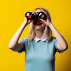 Blonde girl in blue dress watching in binoculars on yellow background