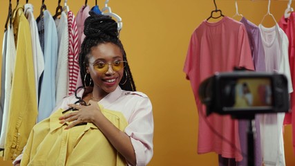 Happy stylish vlogger afro-american girl recording try-on video haul with shopping purchases boasting of clothes on hanger smiling cheerful. Live stream. Fashion concept.