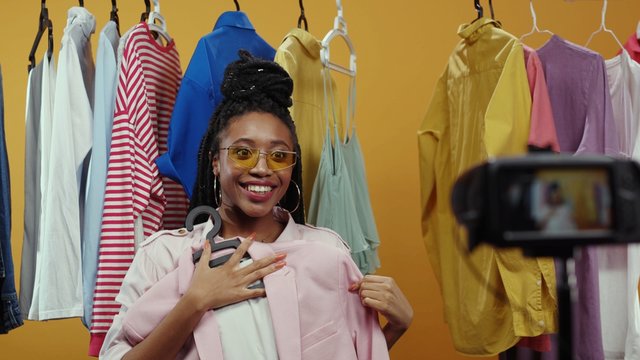 Joyful nice-looking fashion influencer young woman recording live streaming style vlog video talking on camera showing rack of stylish clothes isolated on background.