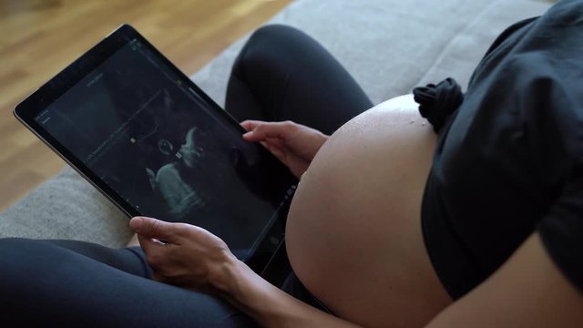 Video of beautiful young pregnant woman looking ultrasound of her baby on digital tablet while lying on bed