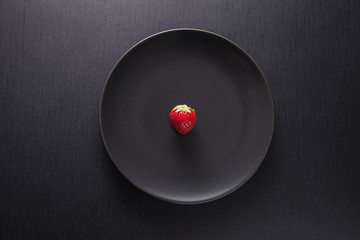 Single red strawberry on a black plate and a black background. Copy space, top view