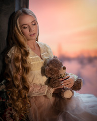 Tender young girl with long hair on a windowsill with a teddy bear