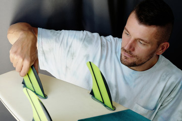 Portrait of a Man attaching Fins on the bottom of his surfboard at home.