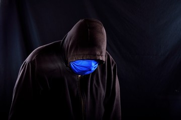 Male figure slouched over wearing black hoodie and blue covid-19 face mask slanted to his left with copy space to the right on a black background. 