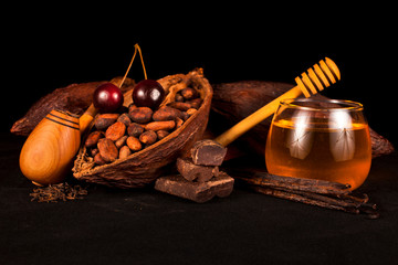 Cocoa pod and cocoa beans, cherry, bourbon vanilla pods, chocolate, tobacco pipe and glass jar of honey. Still life on a black background.