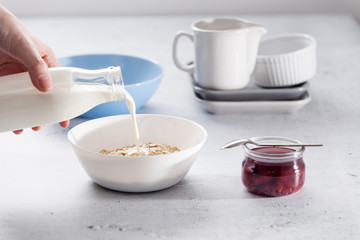 Hand pouring milk into a bowl full of oats on a breakfast table with raspberry jam in a jar, bottle of milk. Copy space, light grey background.