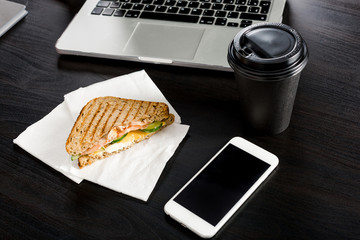 Business lunch concept during the work process. Coffee and a sandwich lying on the office desk next to the phone and laptop.