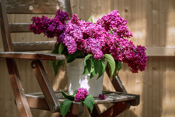A bouquet of lilacs in a white bucket stands on an old garden wooden armchair against the background of a wooden wall.